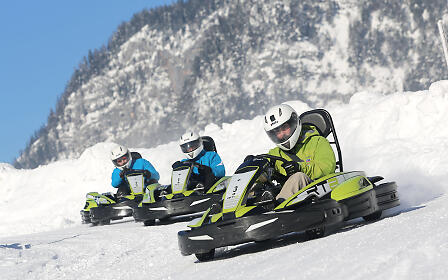 Three people in winter clothing riding snow karts down a snowy slope with a mountainous winter landscape in the background.