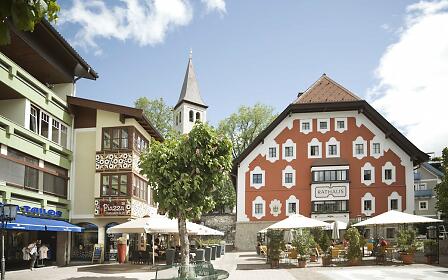 Picturesque village square with historic buildings in warm color tones, surrounded by a church spire and cozy street cafés.