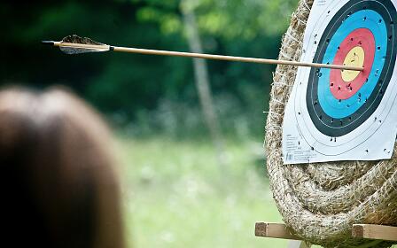 An arrow hits the center of a target during outdoor archery practice. The target is mounted on a straw bale and surrounded by a lush forest backdrop. The precise moment of impact captures focus, skill, and the timeless fascination of archery in the heart of nature.