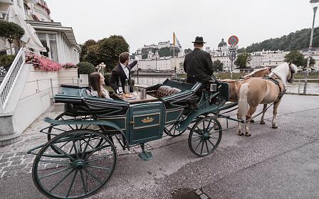 Historic horse-drawn carriage with a beige horse parked on a street corner in Salzburg, with flowering plants and historic buildings in the background.
