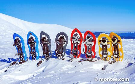 Colorful snowshoes stand lined up in fresh powder snow against a stunning mountain landscape. Beneath a bright blue sky, the vibrant outdoor gear contrasts with the white winter scenery and invites participants to enjoy a shared snowshoe adventure in alpine nature.
