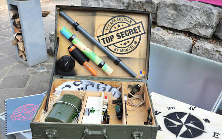 Open military metal case with top secret escape mission equipment including compass, binoculars and navigation materials on stone surface.