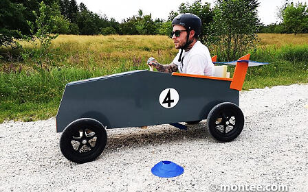 A participant drives a self-built soapbox car along an outdoor course, skillfully maneuvering around marker cones. Wearing a helmet and focused expression, he steers the creative racing vehicle across the gravel path while the natural landscape frames the action-packed team event.