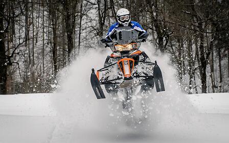 Dynamic winter scene with an orange snowmobile leaping through snow-covered forest landscape, creating snow fountains.