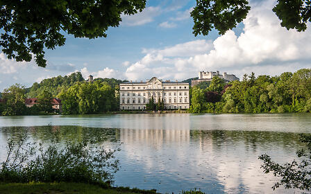 Baroque palace by a lake surrounded by lush greenery, with a white facade and a fortress on a hill in the background.