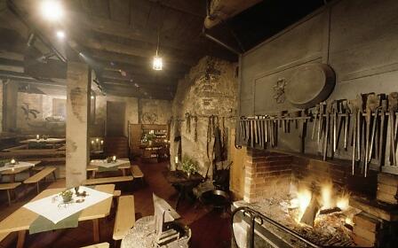 Historische Hammerschmiede mit loderndem Feuer im Kamin, rustikalen Holztischen und hängenden Schmiedewerkzeugen an der Wand.