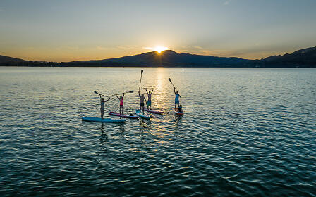 Four stand-up paddleboarders on a calm lake at sunset, framed by gentle mountains and a golden water reflection.