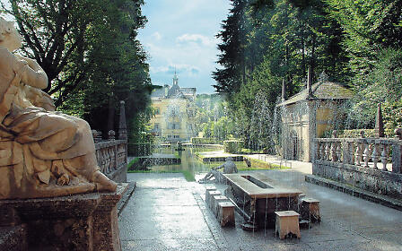 Historical park with stone statue, water features and fountain installation, with a baroque building visible among trees in the background.