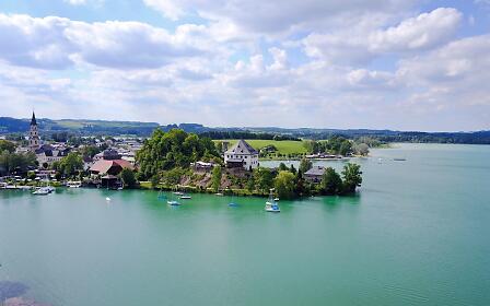 Malerische Seelandschaft mit türkisgrünem Gewässer, weißen Segelschiffen und einer historischen Ortsansiedlung am Ufer, umgeben von sanften Hügeln und bewölktem Sommerhimmel.
