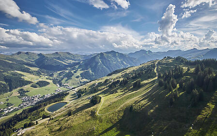 Weite Berglandschaft im Salzburger Land mit grünen Almwiesen, Nadelwäldern und einem kleinen Bergsee. Sanfte Hügel und markante Berggipfel unter einem dramatischen Wolkenhimmel.