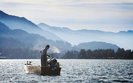 Ein Angler in einem kleinen Ruderboot auf einem glitzernden See, umgeben von nebligen Berglandschaften im SalzburgerLand.