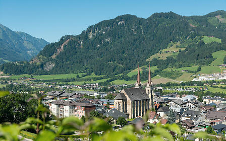 Blick auf eine malerische Alpengemeinde mit gotischer Kirche, umgeben von grünen Wiesen und bewaldeten Berghängen unter strahlend blauem Himmel.