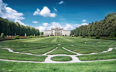 Barockes Schloss mit symmetrischem Barockgarten, grüne Rasenflächen mit kunstvollen weißen Kieswegen und präzise geschnittenen Hecken vor blauem Himmel.