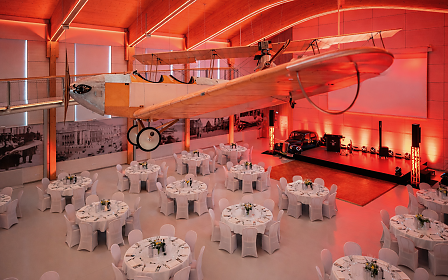 Die außergewöhnliche Eventlocation im fahr(T)raum Mattsee vereint historische Luftfahrt mit stilvoller Veranstaltungsatmosphäre. Unter einem imposanten historischen Flugzeug erstreckt sich ein elegant gedeckter Saal mit runden Tischen, Bühne und eindrucksvoller roter Lichtinszenierung – ideal für Galas, Firmenfeiern, Dinner-Events und exklusive Abendveranstaltungen.