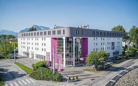 The modern exterior of FourSide Hotel Salzburg with purple highlights and a large glass front stands out. Centrally located with views of the Alps, the hotel offers an attractive setting for both business and leisure.