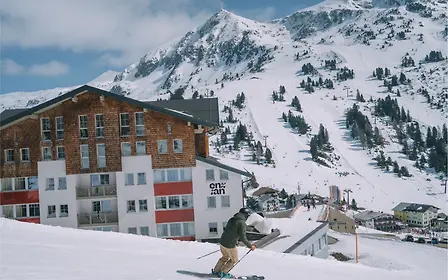 Ein Skifahrer fährt auf der verschneiten Piste unmittelbar am Hotel Enzian Obertauern vorbei. Hinter dem Gebäude erstrecken sich breite Abfahrten, Liftanlagen und markante Berggipfel – ein direkter Blick auf die alpine Lage des Hotels im Salzburger Land.