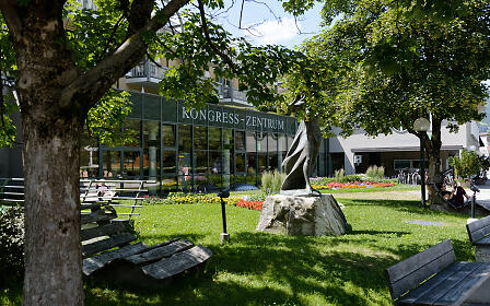 Im sonnendurchfluteten Kurpark von Bad Hofgastein liegt das moderne Kongresszentrum hinter großen Glasfronten. Blumenbeete, alte Bäume und eine stilisierte Bronze-Skulptur schaffen eine einladende Atmosphäre für Gäste und Besucher.
