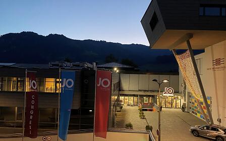 JO CONGRESS in St. Johann glows at dusk. Modern architecture, illuminated entrances and bold flags make a stylish statement against the mountain backdrop.