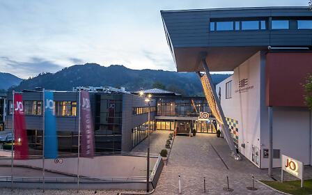 Das JO Congress in St. Johann präsentiert sich in moderner Architektur mit großzügigem Vorplatz, beleuchteten Fassaden und markantem Eingangsbereich. Im Hintergrund schaffen die umliegenden Berge und die Abendstimmung eine repräsentative Kulisse für Events und Kongresse.