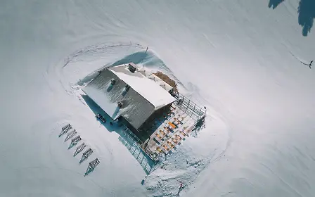 Schrägaufnahme der Mankei Alm inmitten der verschneiten Berglandschaft von Obertauern. Die zum Hotel Enzian gehörende Holzhütte ist von präparierten Skipisten umgeben; auf der sonnigen Terrasse stehen zahlreiche Tische, Bänke und Sonnenschirme für die Gäste bereit.