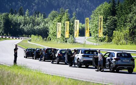Eine Reihe moderner Fahrzeuge hält auf der kurvenreichen Strecke des ÖAMTC Fahrtechnik Zentrums Saalfelden. Teilnehmer steigen aus und bereiten sich auf das Fahrtraining vor, während gelbe Fahnen entlang der Strecke wehen und die grüne alpine Landschaft eine eindrucksvolle Kulisse für das Sicherheitstraining bildet.
