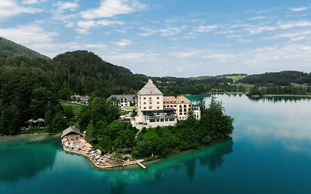Luftaufnahme des exklusiven Rosewood Schloss Fuschl direkt am See. Im Hintergrund blauer Himmel und traumhafte Berglandschaft und im Vordergrund spiegelt der See