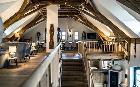 Der offene Galeriebereich der Remise beeindruckt mit massiven Holzbalken, gemütlichen Sitzgruppen und alpiner Dekoration. Die Kombination aus rustikalem Charme und stilvoller Einrichtung bietet einen besonderen Rahmen für Events und entspannte Zusammenkünfte.