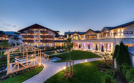 Stimmungsvolle Abendaufnahme eines modernen Wellnesshotels mit beleuchtetem Garten, stilvollen Liegeplätzen und offenem Spa-Bereich. Warme Lichtakzente verleihen dem alpinen Gebäudeensemble einladende Eleganz und Wohlfühlcharakter.