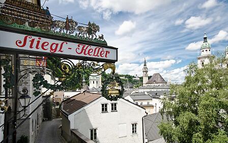 The ornate Stiegl-Keller sign with a brewery wagon motif marks the entrance to this traditional Salzburg tavern. Behind it, the historic Old Town, cathedral towers and Hohensalzburg Fortress rise under a vibrant sky – a beloved panoramic spot.