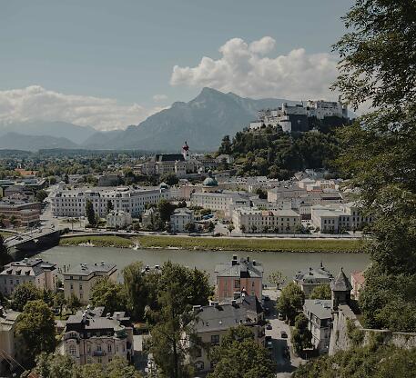 Blick auf die historische Altstadt Salzburgs mit der Festung Hohensalzburg, eingebettet zwischen Bergen und der Salzach, unter wolkigem Sommerhimmel.