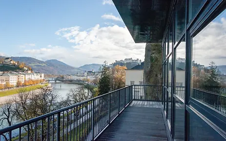 Moderner Balkon mit Blick auf die Salzach und die historische Altstadt Salzburgs, umrahmt von herbstlichen Bäumen und Bergen.
