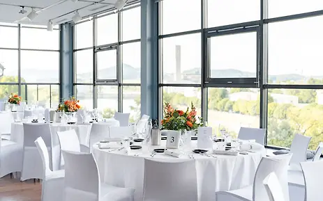 Eleganter Veranstaltungsraum mit großen Glasfenstern und panoramischen Ausblicken, gedeckt mit weißen Tischen und orangefarbenen Blumenarrangements.