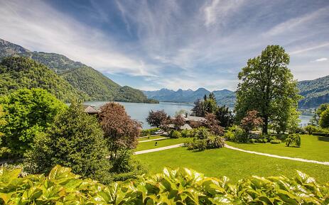 Amidst a breathtaking natural backdrop, a carefully designed park unfolds with gentle paths winding through lush green lawns. In the background, a crystal-clear mountain lake stretches out, framed by majestic mountain ranges whose forested slopes gently rise into the azure blue sky. Picturesque park landscape overlooking a mountain lake, surrounded by green hills and forested mountain ranges under a dynamic cloudy sky.