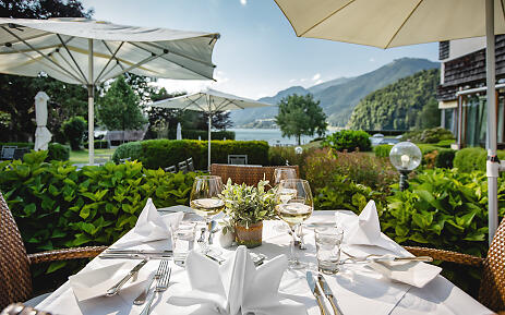 Amidst a breathtaking mountain landscape, a carefully set table invites you to indulge. White linen napkins, elegant wine glasses, and a delicate plant decoration frame a culinary experience at Seehotel Brandauer's Villen, where nature and gastronomy merge in perfect harmony. Outdoor table setting with a view of a picturesque mountain lake, surrounded by lush gardens and white umbrellas.