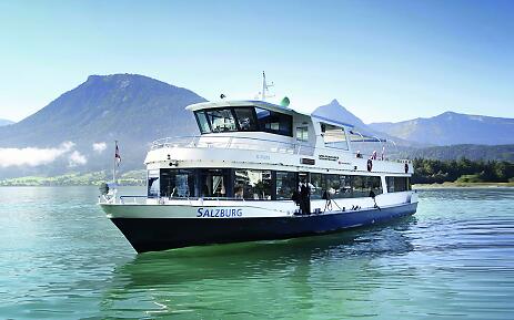 Amidst a breathtaking mountain landscape, an elegant excursion boat glides across the crystal-clear waters. Soft mountain silhouettes and a bright blue sky frame the majestic scenery, while the ship embarks on a tranquil journey through the impressive natural backdrop. White excursion boat on a turquoise lake with mountains in the background, part of the Salzburg AG fleet.