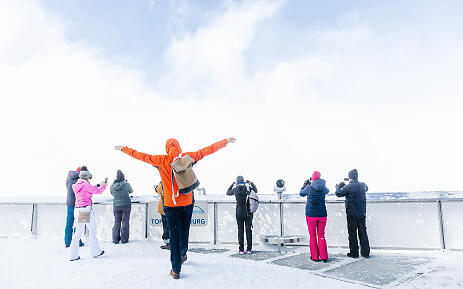 Auf einer verschneiten Aussichtsplattform stehen Besucher in warmer Winterkleidung und genießen die atemberaubende Weite der winterlichen Berglandschaft. Eine Person in orangefarbener Jacke breitet die Arme aus und feiert die pure Freiheit und Schönheit des Moments. Winterliche Aussichtsplattform mit Besuchern in bunten Jacken, die die verschneite Berglandschaft betrachten und fotografieren.