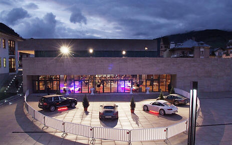 Abendliche Präsentation von Luxusfahrzeugen vor einem modernen Gebäude mit lila-blauer Beleuchtung, umgeben von Bergen und nächtlichem Himmel.