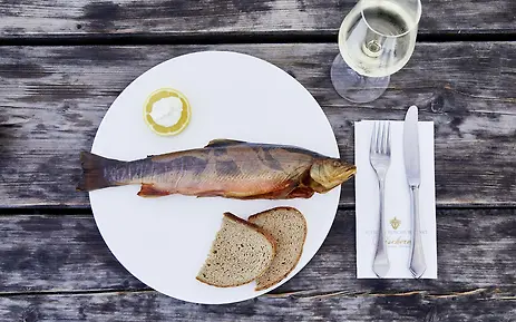 Fresh prepared fish on white plate, served with bread slices and lemon on rustic wooden surface, accompanied by a white wine glass.