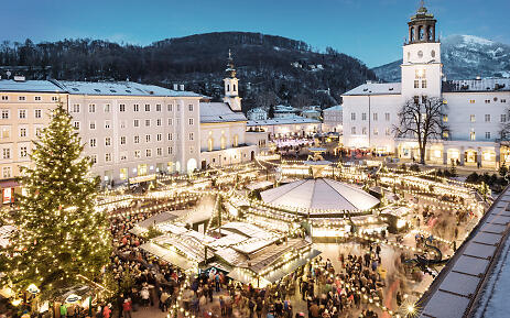 In the snowy backdrop of Salzburg, a magical Christmas market unfolds that makes the heart skip a beat. Hundreds of lights bathe the square in warm, golden light, while numerous market stalls are invitingly arranged between historic buildings. The surrounding mountains and baroque architecture lend the market a fairytale-like atmosphere. Christmas market in historic Salzburg old town setting with festively illuminated market stalls, large Christmas tree, and snow-covered buildings against a winter mountain landscape.