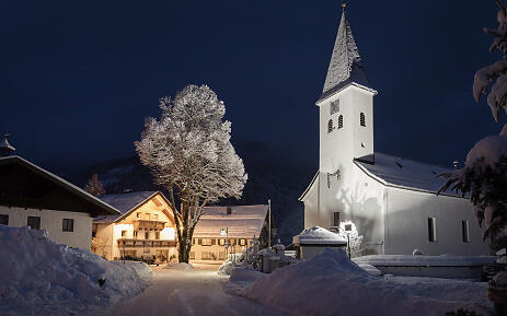 A magical winter landscape reveals itself in nocturnal silence. The white village church with its slender tower stands majestically next to a snow-covered farmhouse, while an illuminated tree bathes the scenery in a warm, inviting light. High snow banks line the path, testifying to an intense winter in this idyllic mountain region. Nighttime winter village scene with snow-covered church and warmly lit buildings, nestled in a snowy landscape with an illuminated tree.