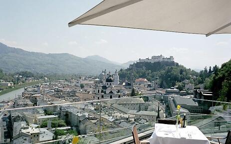 From a sun-drenched panoramic terrace, the view sweeps over the densely built old town of Salzburg. In the background, the white Hohensalzburg Fortress sits majestically on a wooded hill, while baroque church domes and historic buildings harmoniously blend into the cityscape. The gentle mountains of the Salzburg region frame this breathtaking backdrop. View from a terrace overlooking the historic old town of Salzburg with Hohensalzburg Fortress, surrounded by forested mountains and a picturesque river landscape.