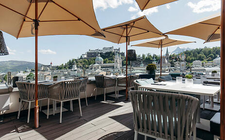 An elegant rooftop terrace invites guests to linger, with artfully woven chairs and white tables. Warm beige sunshades provide shade and frame the breathtaking view of Salzburg's picturesque cityscape. In the background, the majestic Hohensalzburg Fortress towers over the historic rooftops and church spires of the city. Sun-drenched rooftop terrace with shaded umbrellas offering a panoramic view of Salzburg's historic old town and Hohensalzburg Fortress.