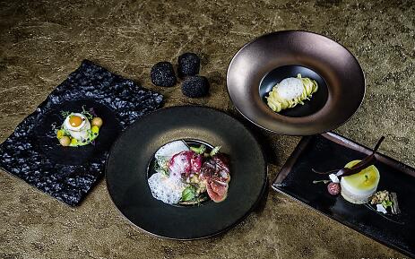 Ein kunstvoll komponiertes Gourmet-Menü entfaltet sich auf edlem Geschirr: zartes Fleisch mit Schaum, feine Pasta, ein raffiniertes Dessert und filigrane Vorspeise – begleitet von schwarzen Trüffeln auf samtigem Untergrund in luxuriösem Ambiente. Mehrgängiges Gourmet-Menü mit Fleisch, Pasta, Dessert und Trüffeln kunstvoll angerichtet.