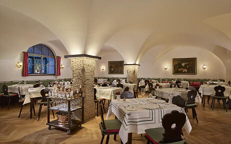 At Hotel Goldener Hirsch in Salzburg, historic vaulted ceilings meet elegant interiors. Rustic stone columns, classic wooden chairs, and finely set tables create a stylish atmosphere full of Salzburg’s authenticity and history. Hotel Goldener Hirsch in Salzburg with vaulted ceiling, stone columns, and set tables.