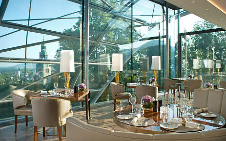 Ein elegantes Restaurant mit Panoramaverglasung eröffnet den Blick auf eine malerische Stadt und grüne Baumkronen. Goldene Akzente, edle Materialien und stilvoll gedeckte Tische schaffen ein luxuriöses Ambiente in lichtdurchfluteter Architektur. Modernes Luxusrestaurant mit Glasfront, Ausblick, goldenen Details und gedeckten Tischen.