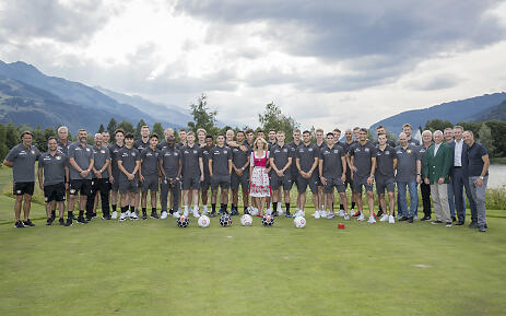 Large football team in gray jerseys poses on a meadow with mountains in the background, surrounded by several footballs.