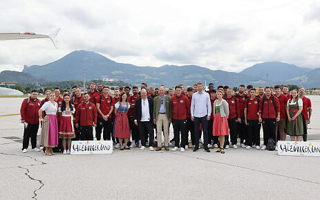 Against the majestic mountain backdrop of SalzburgerLand, an impressive group of athletes and honored guests gather. The protagonists in bright red jackets stand confidently on the airfield, framed by gentle mountain ranges and wide meadows - a moment of connection and regional identity. Large group photo on an airfield with mountain panorama, consisting of athletes in red jackets and some people in traditional costume.