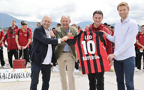 In the picturesque mountain landscape of SalzburgerLand, a festive atmosphere emerges: sports officials and team representatives celebrate a special moment with the presentation of a personalized jersey. In the background, players in uniform red jackets look on, while gentle mountains line the horizon. Four men display a Bayer 04 Leverkusen jersey against a mountain backdrop, surrounded by football players in red jackets.