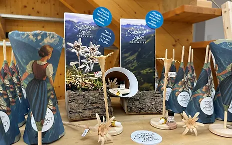 In einer warmen Holzatmosphäre präsentieren sich die handgefertigten „Original Salzburger Zirben-Edelweiss“-Produkte auf stilvollen Holzsockeln. Naturmaterialien, Edelweiß-Blüten und alpine Motive unterstreichen die hochwertige, traditionelle Handwerkskunst und den authentischen Duft der Alpenwelt.