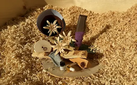Eingebettet in feine Zirbenspäne liegt eine kunstvolle Auswahl an handgefertigten Holzobjekten, Edelweiß-Blüten und naturbelassenen Duftaccessoires. Das stimmungsvolle Licht hebt die alpine Ästhetik und die traditionelle Verarbeitung der Salzburger Zirbenprodukte hervor.